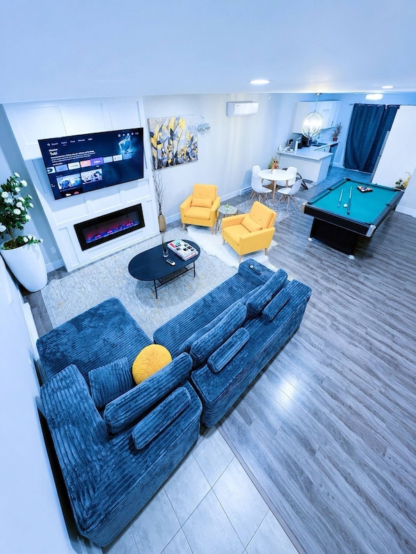 A modern living room with a blue sofa, yellow armchairs, a pool table, and a flat-screen TV mounted on the wall. - Modern 3-bedroom Retreat Near Moncton Hospital and Downtown (Moncton)