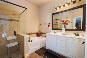 Ein Badezimmer mit Duschkabine aus Glas, einer weißen Badewanne, einem Waschtisch mit großem Spiegel und einer roten Blumenvase.