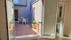 Une pièce dotée d’une porte-fenêtre coulissante donnant sur un patio extérieur avec une table et des chaises, des plantes en pot et une vue sur un immeuble avec un balcon.