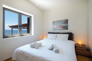 A bedroom with a large window, a bed with white linens, two bedside lamps, and a painting on the wall. - Villa Isabelle on the beach (Iria)