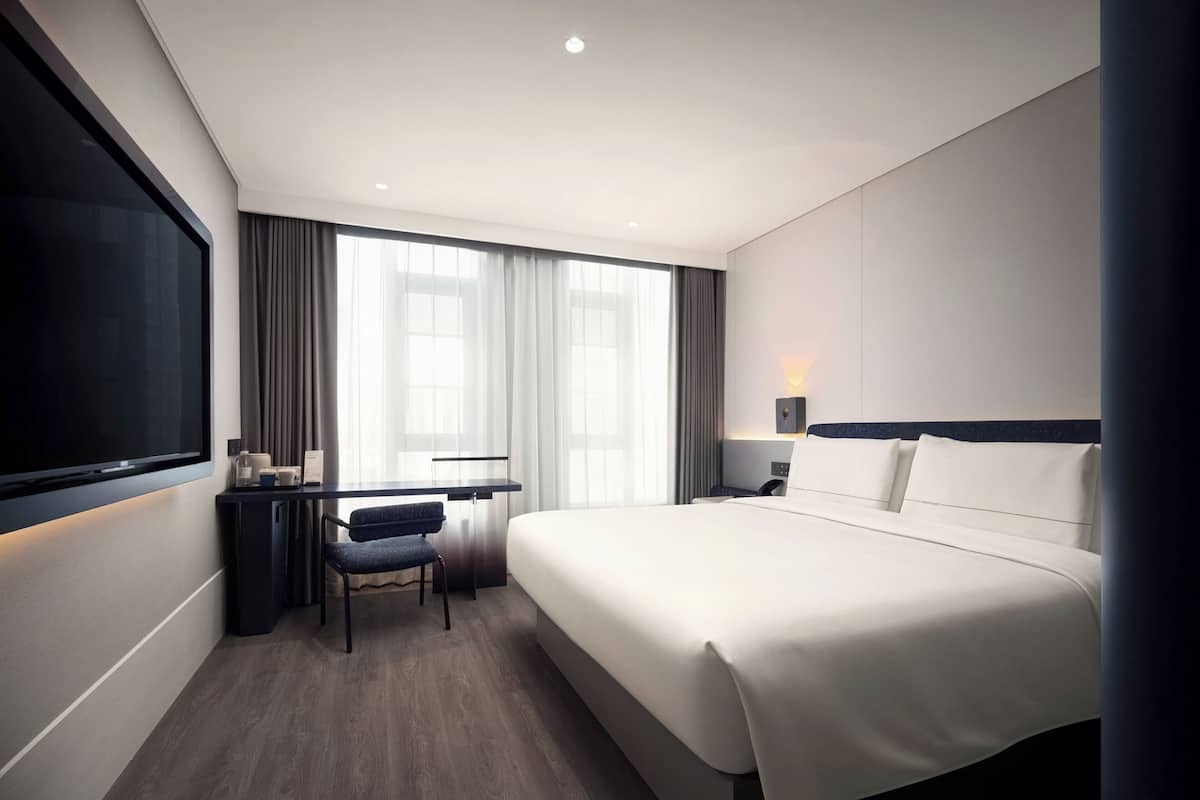 Habitación de hotel con una cama grande, un escritorio con silla, un televisor de pantalla plana y una ventana con cortinas.