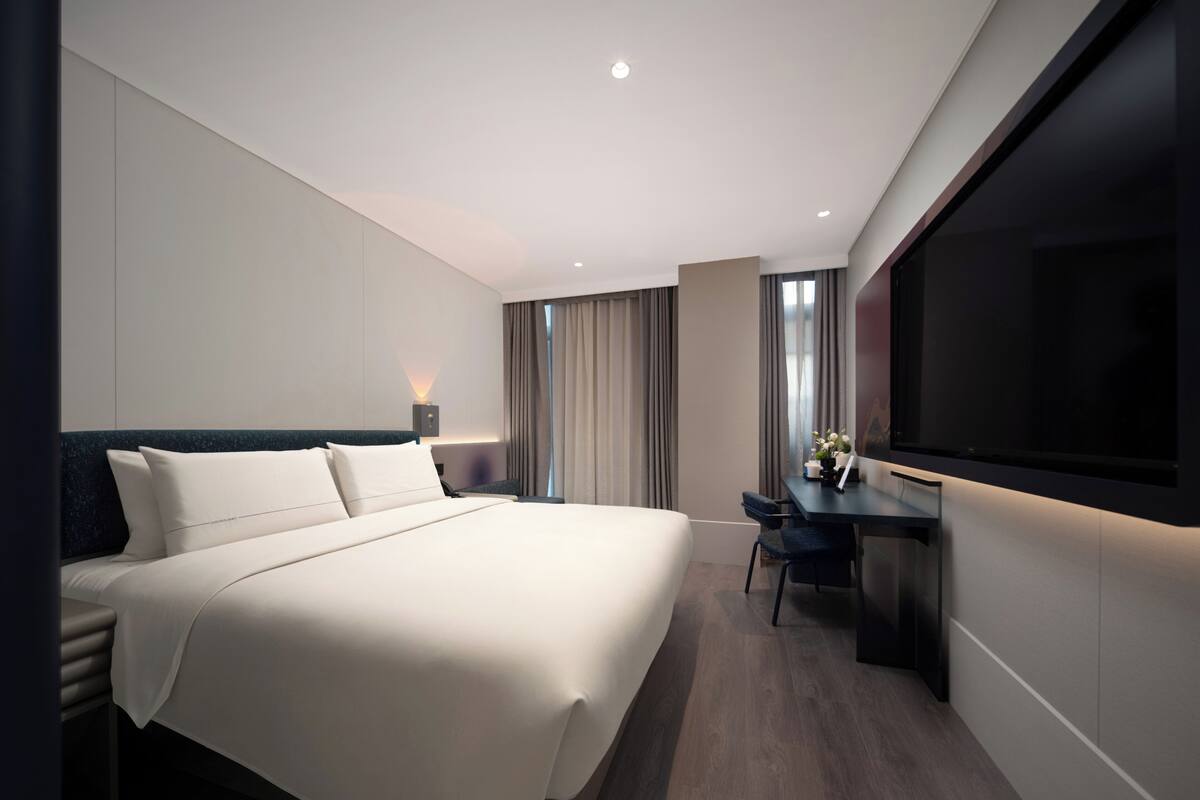 Habitación de hotel moderna con una cama grande, un escritorio, un televisor de pantalla plana y un ventanal con cortinas.
