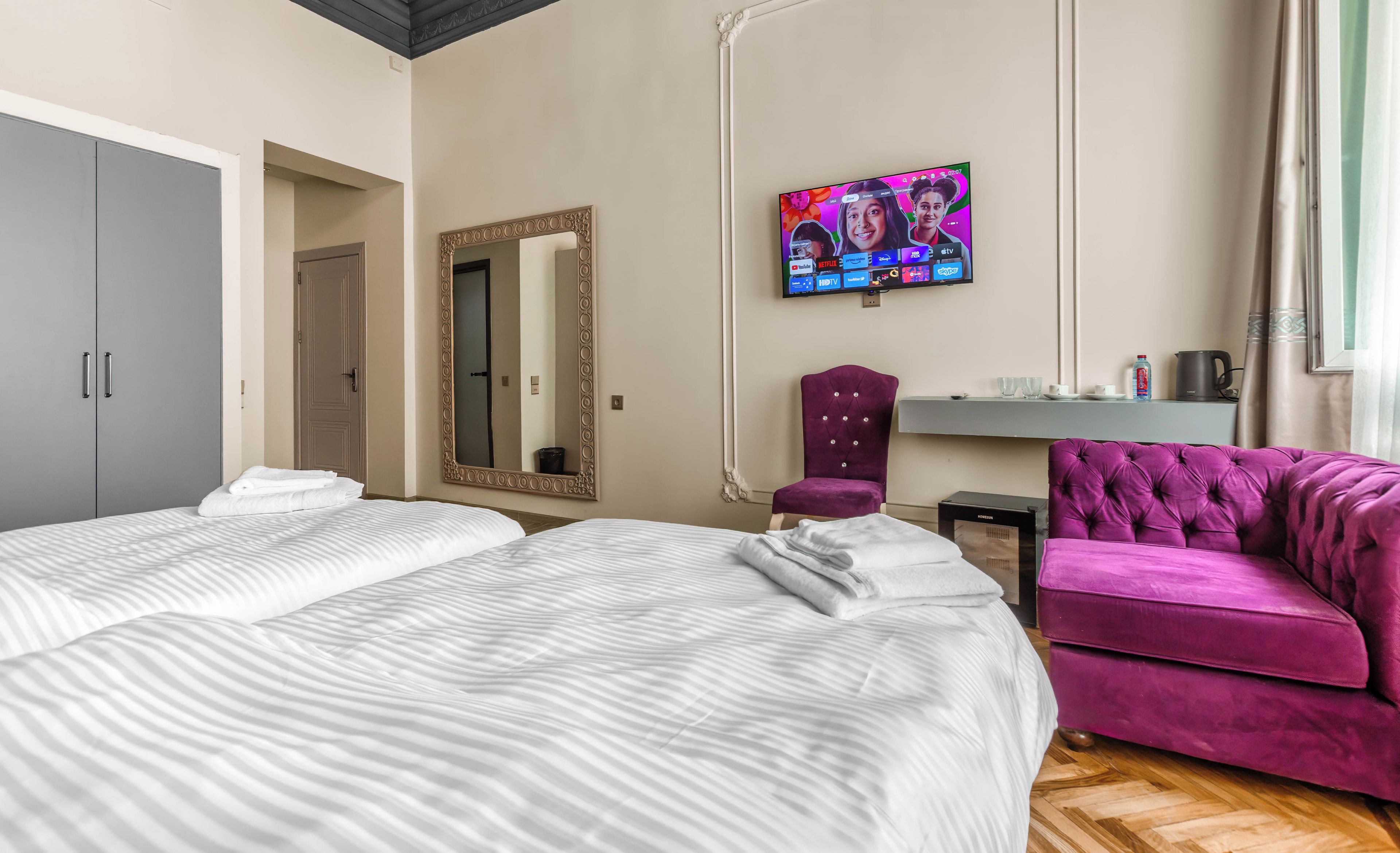 Habitació d'hotel amb dos llits, un mirall gran, televisió i una butaca morada.