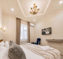 Een hotelkamer met een groot bed, een kroonluchter, een raam met gordijnen, een stoel, een schilderij aan de muur en een plank met bekers en glazen.