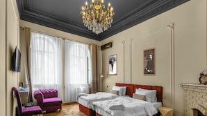 Ett hotellrum med två sängar, en öppen spis, en kristallkrona och ett stort fönster med gardiner.