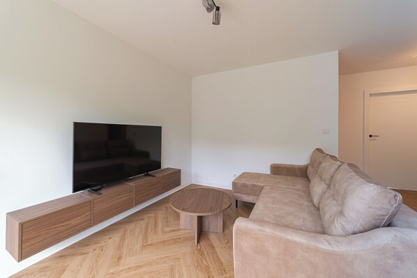 Ein modernes Wohnzimmer mit Flachbildfernseher, einer Holzkonsole, einem runden Couchtisch und einem beigefarbenen Sofa.