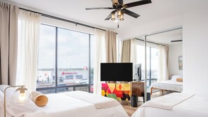 Quarto de hotel com duas camas, televisão, ventoinha no teto e janelas amplas com vista para a cidade.