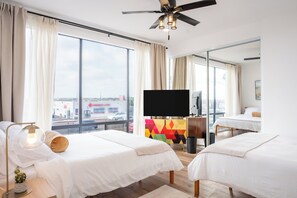 A hotel room with two beds, a TV, a ceiling fan, and large windows offering a city view.