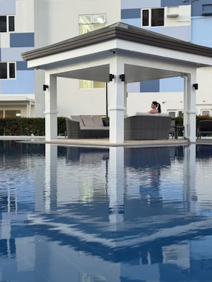 Un área de piscina moderna al aire libre con una zona de estar techada y una persona relajándose.