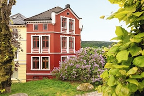 A red building with white trim, a tree, and purple flowers. - Sentrum Suites (Vestland)