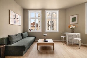 A living room with a sofa, a coffee table, a desk with two chairs, and two windows with a view of buildings.