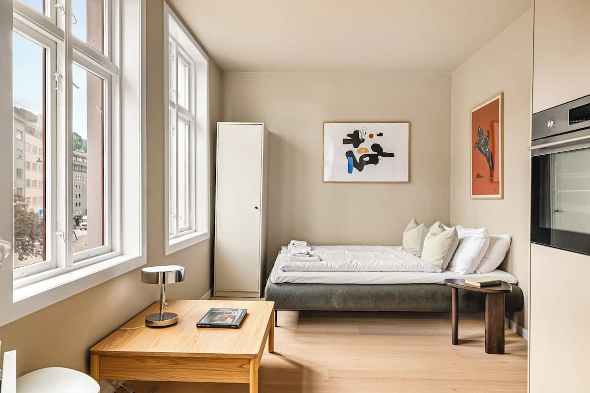 A modern bedroom with a bed, bedside table, lamp, artwork, and a view of the city through large windows.