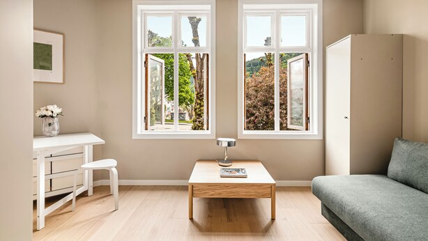 A room with a desk, chair, table, sofa, and two windows with a view of greenery. - Sentrum Suites (Vestland)
