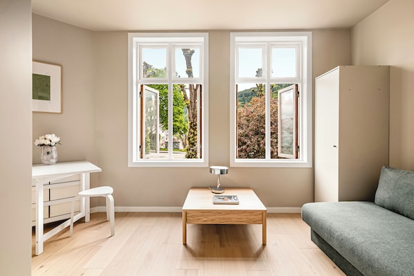 A room with a desk, chair, table, sofa, and two windows with a view of greenery.