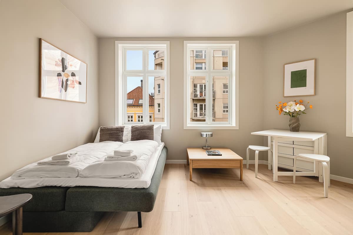A modern bedroom with a bed, bedside tables, a desk, and a view of buildings through large windows.