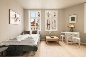 A modern bedroom with a bed, bedside tables, a desk, and a view of buildings through large windows.
