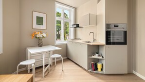 A compact kitchen with a white countertop, a sink, and an oven. There is a window providing natural light, and a small white table with two stools. - Sentrum Suites (Vestland)
