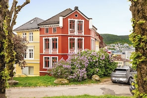 A two-story building with a red and yellow facade, a parked car, and a hill with houses in the background.