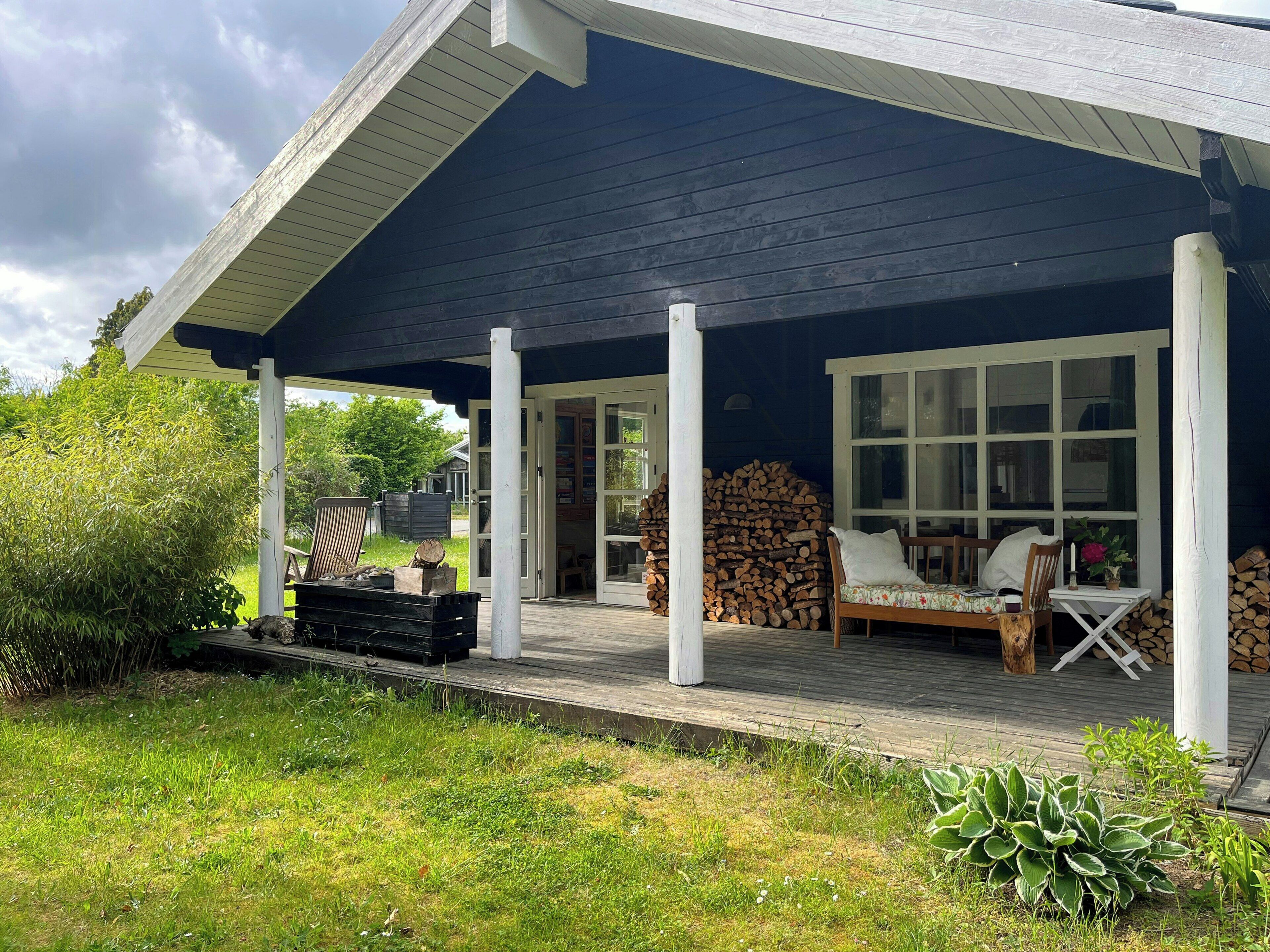A covered outdoor seating area with a wooden bench and a table, surrounded by a garden with a variety of plants.