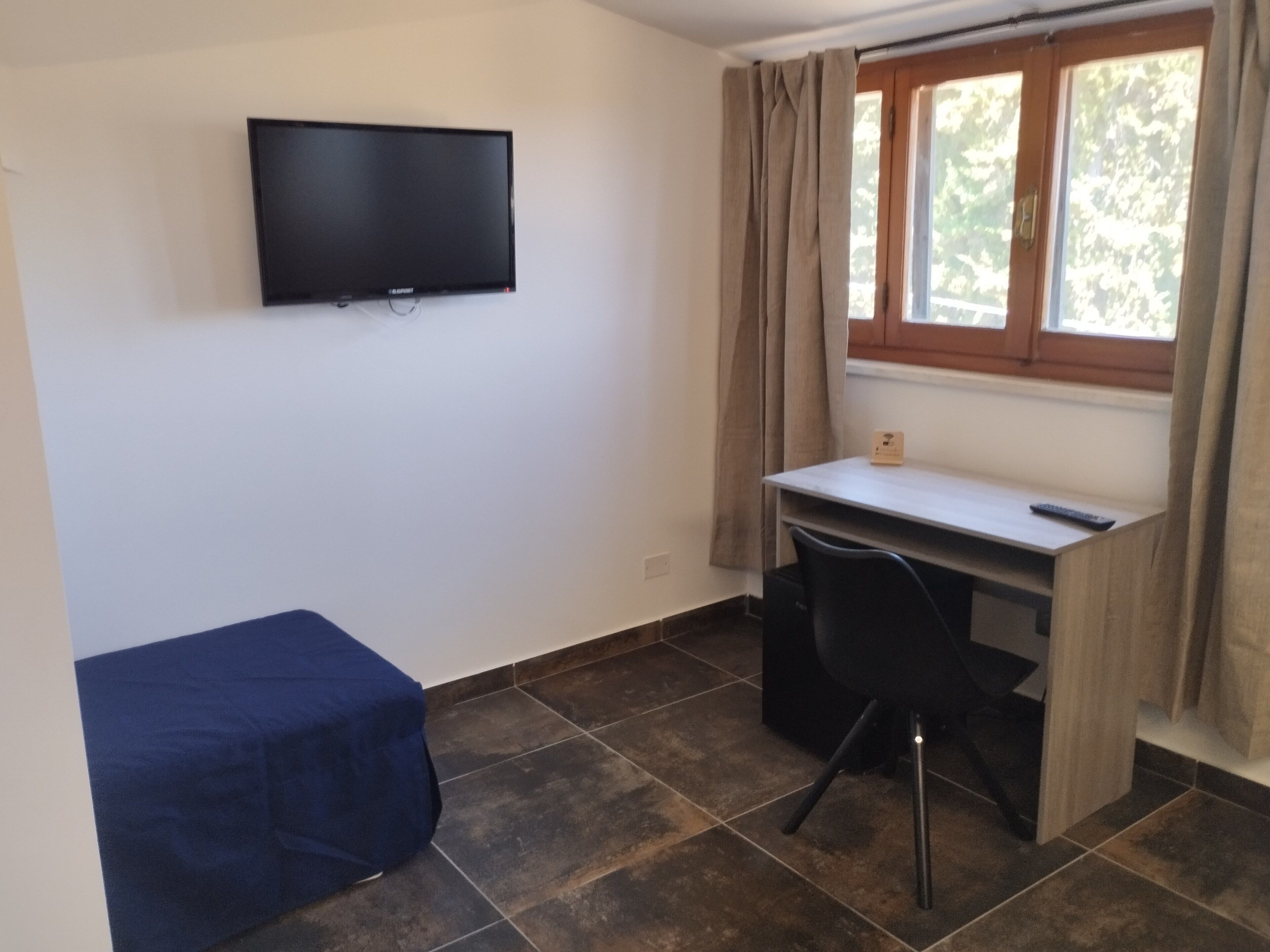 A small room with a bed, a desk, a chair, a TV mounted on the wall, and a window with curtains.