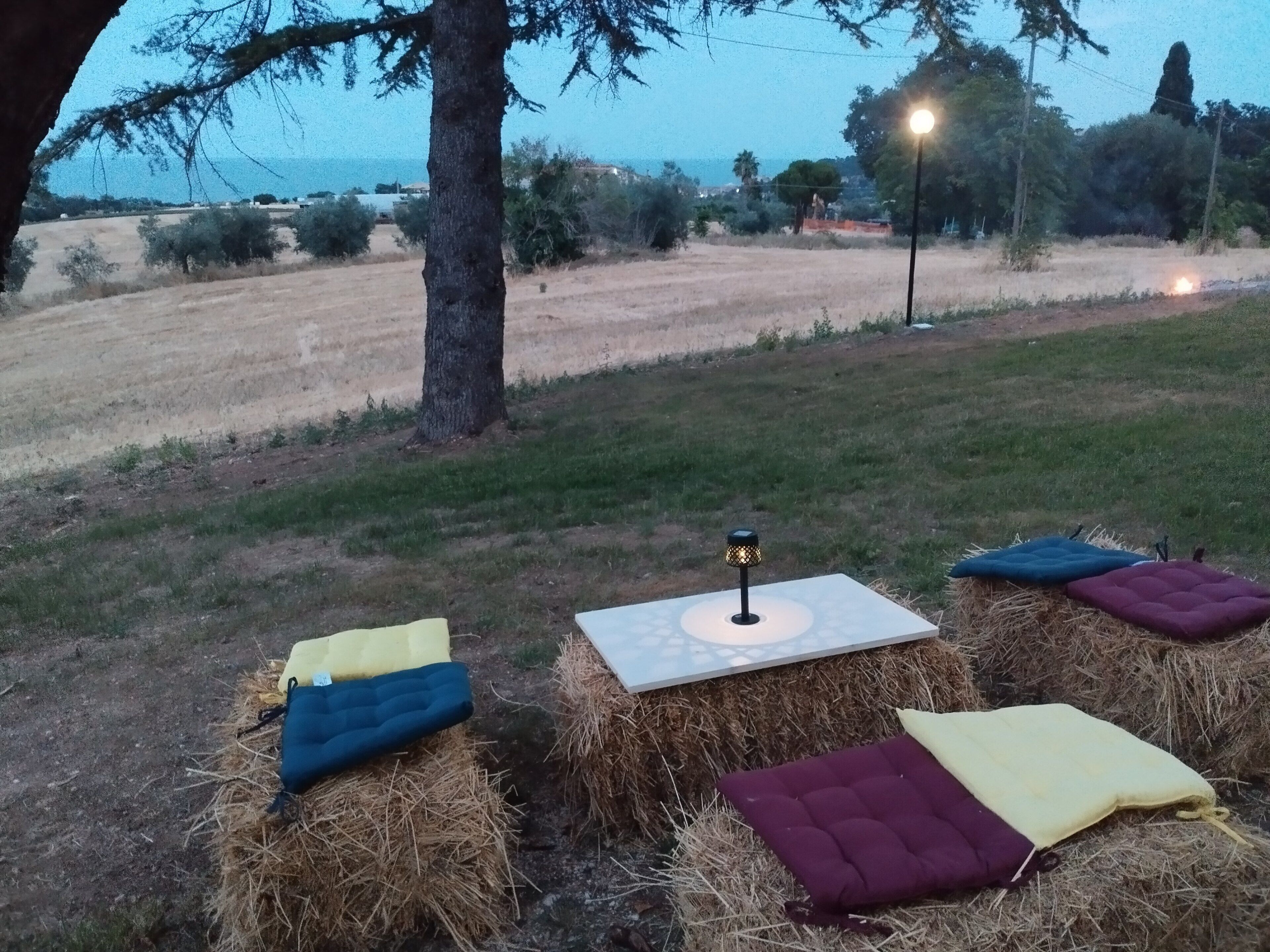 A rustic outdoor seating area with hay bales and cushions, a table with a lamp, and a view of a field and trees.