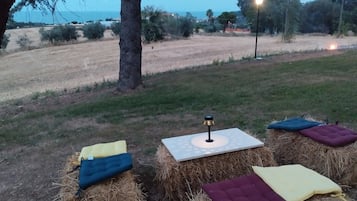 A rustic outdoor seating area with hay bales and cushions, a table with a lamp, and a view of a field and trees.