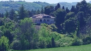 Ein Haus mit geneigtem Dach, umgeben von dichtem Grün und Hügeln im Hintergrund.