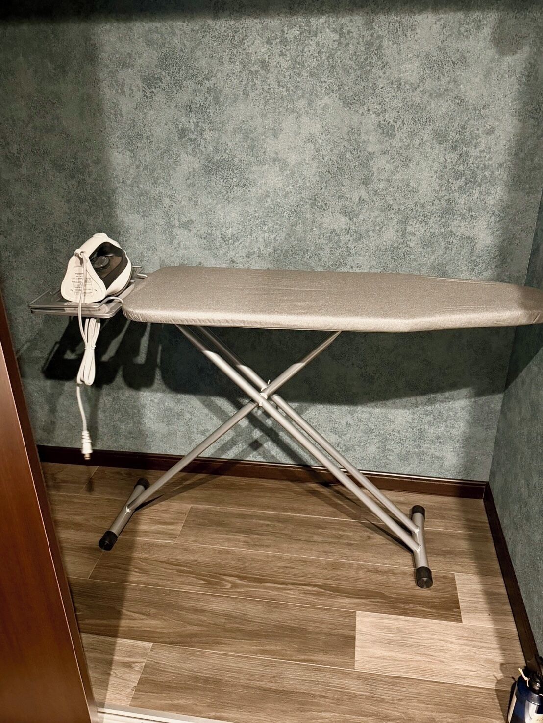 An ironing board with a corded iron, a wooden floor, and a textured wall.