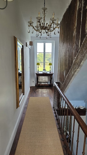 A hallway with a wooden floor, a chandelier, a mirror, a table with lamps, and a staircase. - Charming house nestled in peaceful, authentic surroundings. (Ars)