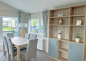A room with a dining table, chairs, a lamp, a radiator, a window with curtains, and a wooden bookshelf with items on it. - Clearwater Pet Friendly (No Hot Tub) (Hexham)