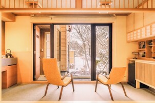 Habitación con dos sillas, un escritorio y vistas a un espacio exterior con una banca y árboles a través de una puerta de vidrio.