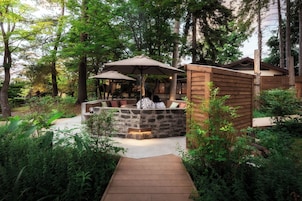 A stone bench with cushions, surrounded by greenery, and a wooden structure with an umbrella.