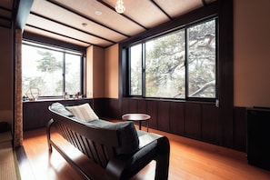Ein Zimmer mit einer hölzernen Sitzbank, einem kleinen runden Tisch und großen Fenstern mit Blick auf Bäume.