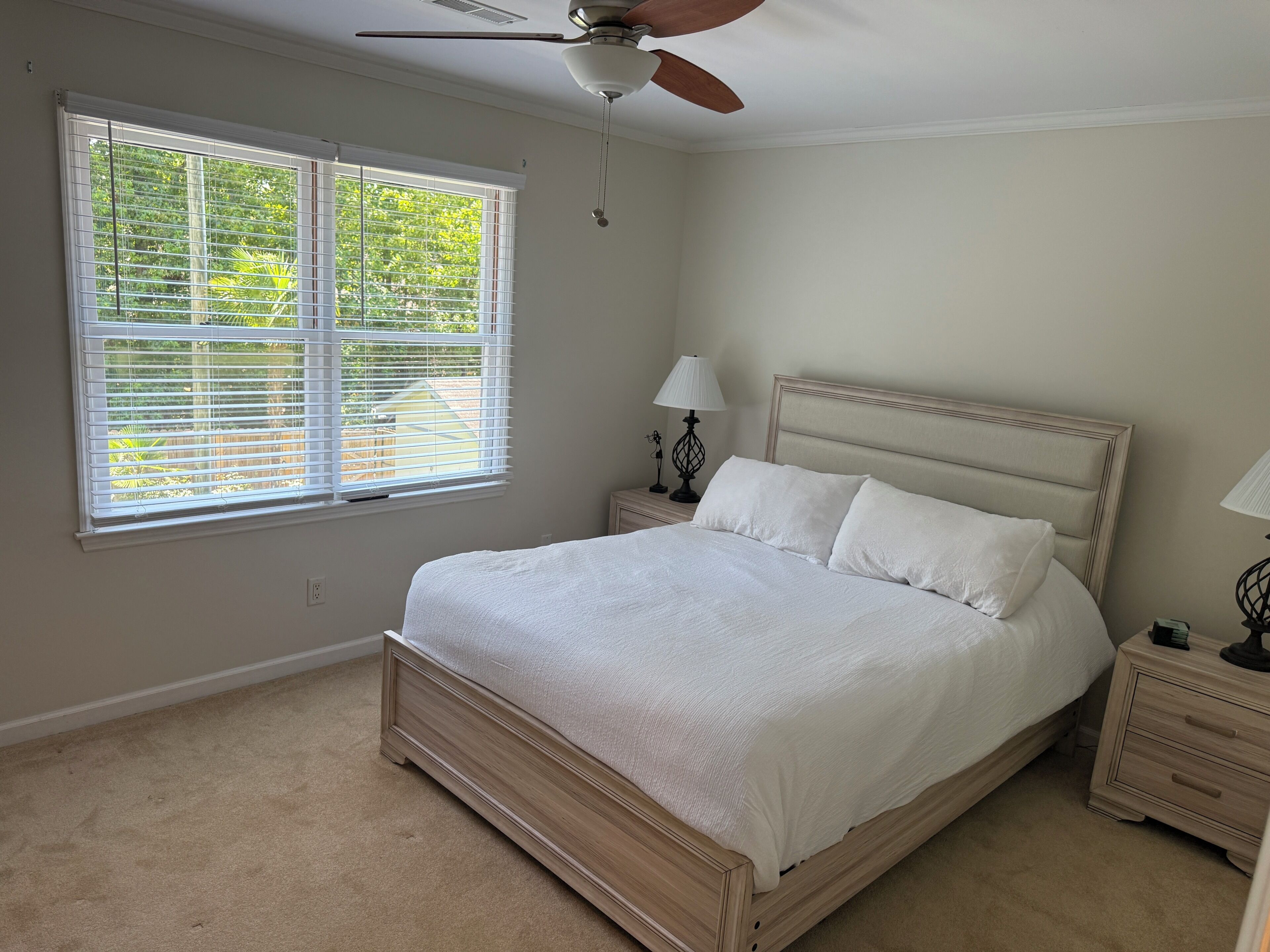 A bedroom with a bed, a ceiling fan, a window with blinds, and a nightstand with a lamp.