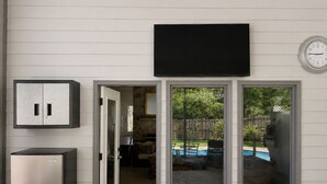 Une maison dotée d’une porte-fenêtre coulissante donnant sur un jardin avec une piscine, un téléviseur fixé au mur et un réfrigérateur.