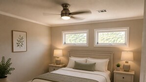 Une chambre à coucher comprenant un lit, deux tables de chevet, un ventilateur de plafond, deux fenêtres, une plante en pot, un miroir et une porte.