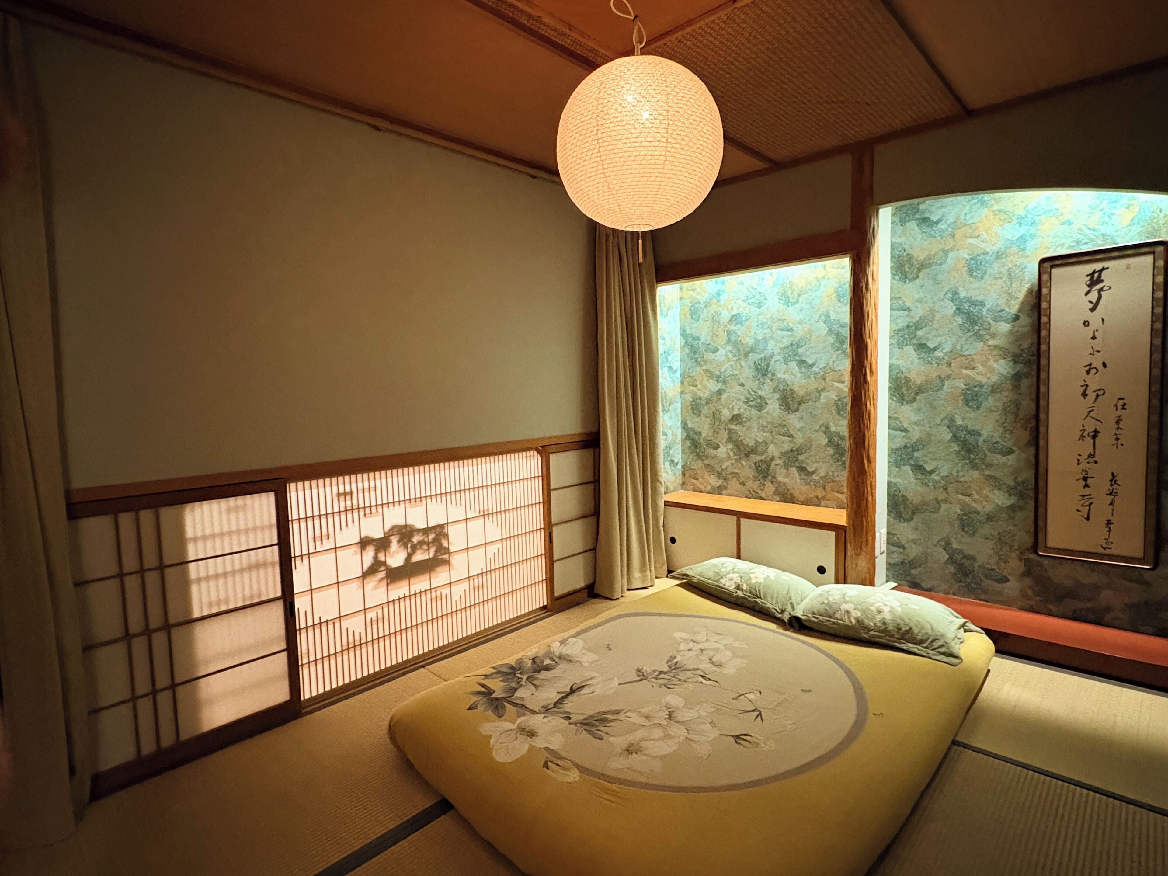 A traditional Japanese room with tatami flooring, a low bed with a floral bedspread, a hanging paper lantern, and a shoji screen with a floral pattern.