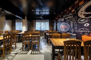 Un restaurante con mesas y sillas de madera, una pared blanco y negro con gráficos relacionados al café, y amplias ventanas con cortinas.