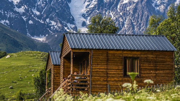 Una cabaña de madera con techo de metal, rodeada por un campo verde exuberante y montañas al fondo.