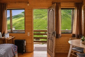 A wooden cabin with a bed, a table, and a view of green hills. - Ushguli cabins (Mestia)