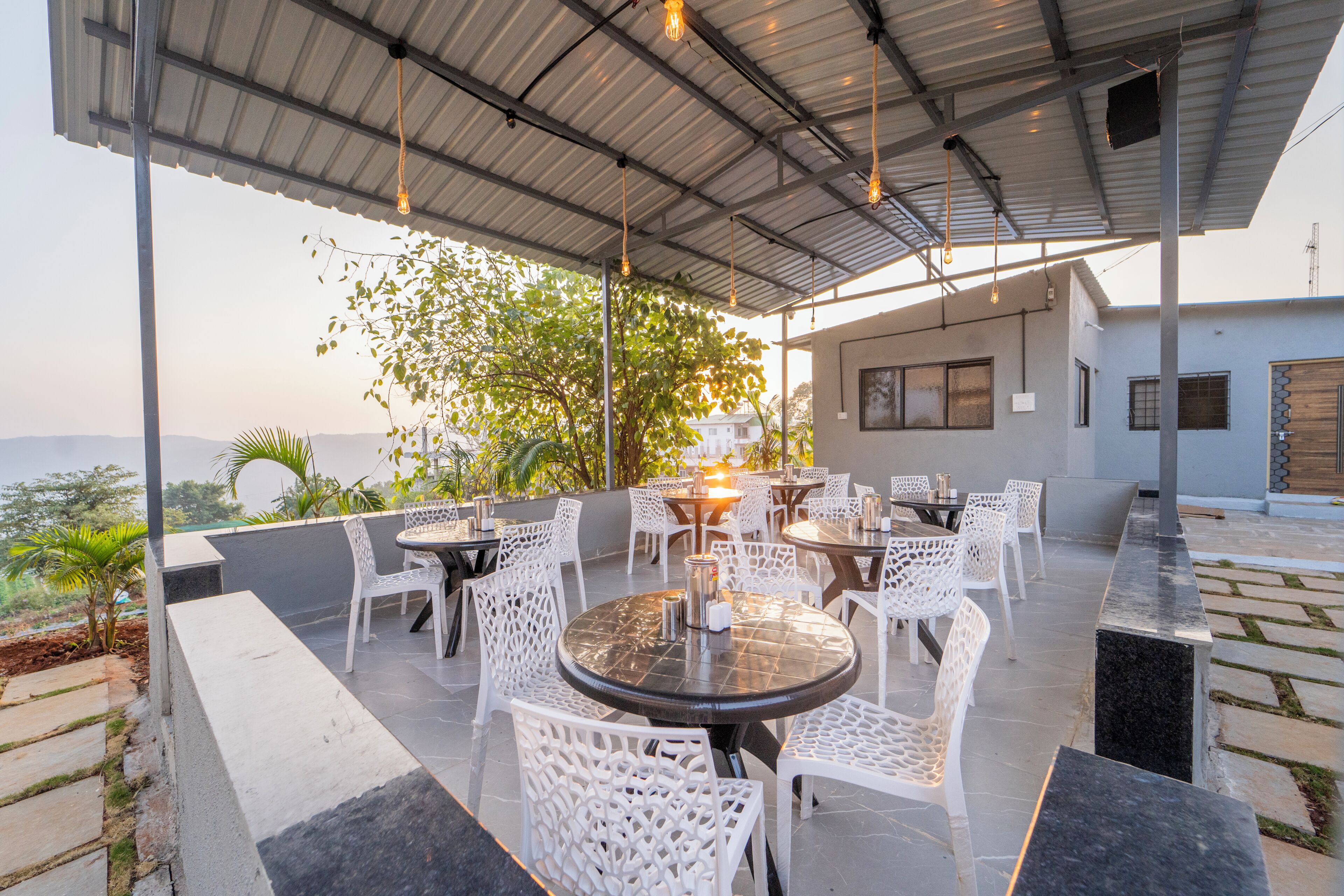 Un espace de restauration en plein air avec des tables rondes et des chaises blanches, entouré d’une terrasse couverte offrant une vue sur le paysage.