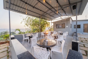 Un espace de restauration en plein air avec des tables rondes et des chaises blanches, entouré d’une terrasse couverte offrant une vue sur le paysage.
