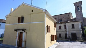 Um edifício de dois andares com fachada amarela, uma pequena entrada com porta, e uma torre de sino ao fundo.