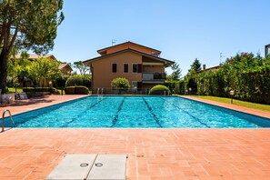 A swimming pool with a brick deck, a two-story building with a balcony in the background, and a clear blue sky.