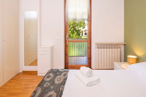 Un dormitorio con piso de madera, una cama con ropa blanca, un plaid estampado, un radiador, una mesita de noche con lámpara, un armario y una puerta que da a un balcón con vistas a la vegetación.