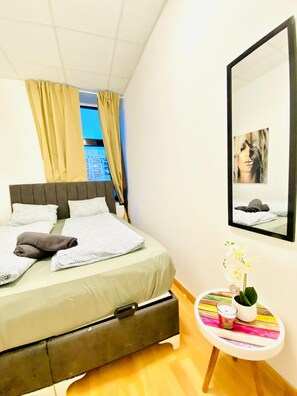 A neatly made bed with a grey headboard, a small bedside table with a vase of flowers, and a large mirror on the wall. - Private Room Long Stay Shared Apt (Wien)