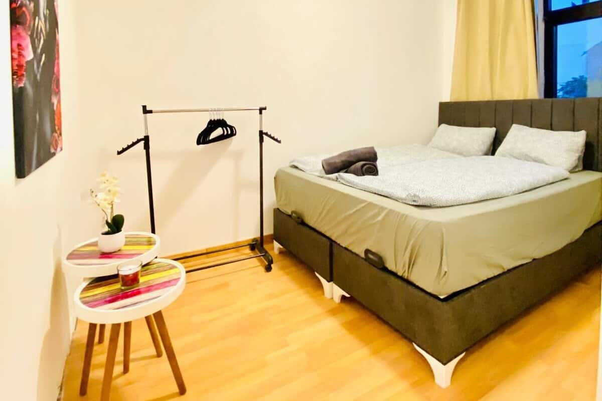 A bedroom with a bed, a small table with a plant, a hanging rack, and a window with curtains.