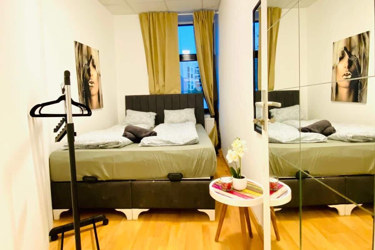 A hotel room with two beds, a standing mirror, a small table with a vase, and a window with curtains.