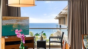 Ein Zimmer mit einem großen Fernseher, einem Holzschrank, einem Tisch mit Stühlen, einer Topfpflanze und Blick aufs Meer.