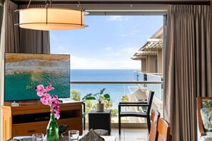 Ein Zimmer mit einem großen Fernseher, einem Holzschrank, einem Tisch mit Stühlen, einer Topfpflanze und Blick aufs Meer.
